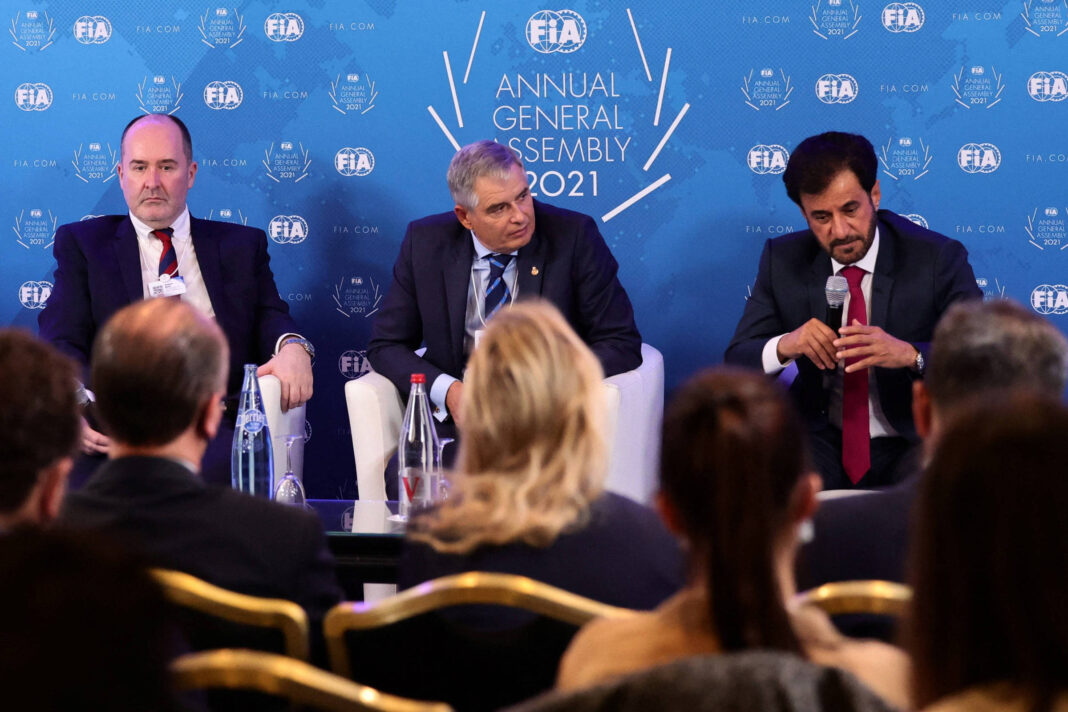 A imagem mostra um painel de discussão durante a Assembleia Geral Anual da FIA em 2021. Três homens estão sentados em cadeiras brancas, com um fundo azul que exibe o logotipo da FIA. O homem à esquerda está vestido com um terno escuro e uma gravata, o do meio usa um terno claro e o da direita está com um terno escuro e uma gravata vermelha. O público é visível ao fundo, com algumas pessoas de costas.
