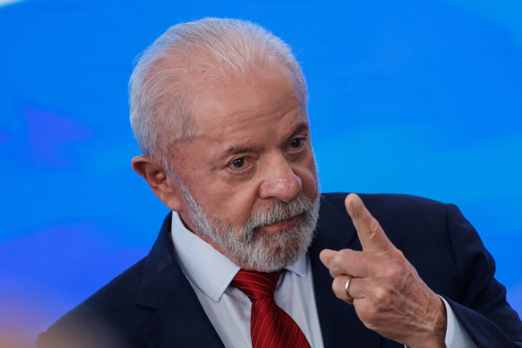 Um homem de cabelo grisalho e barba, vestindo um terno escuro e uma gravata vermelha, está gesticulando com a mão direita levantada e o dedo indicador apontado. Ele parece estar em um ambiente formal, com um fundo azul desfocado.