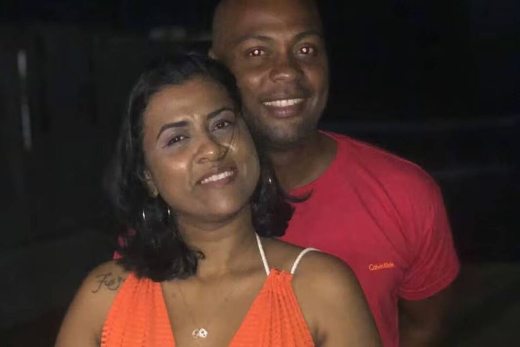 A imagem mostra um casal posando juntos em um ambiente noturno. A mulher está usando uma blusa laranja e tem cabelo escuro e liso, enquanto o homem está usando uma camiseta vermelha. Ambos estão sorrindo e parecem estar felizes. O fundo é escuro, sugerindo que a foto foi tirada à noite.