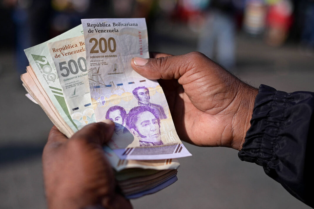 Uma mão segurando várias notas de dinheiro venezuelano. As notas visíveis incluem uma de 200 bolívares e uma de 500 bolívares. O fundo é desfocado, mostrando pessoas ao longe.