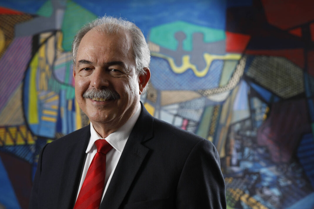 Um homem de cabelos grisalhos e bigode, vestindo um terno escuro e uma gravata vermelha, está posando para a foto. Ele sorri levemente e está em frente a um fundo colorido com formas abstratas e traços artísticos. O fundo apresenta uma combinação de cores como azul, vermelho e amarelo, com elementos geométricos e linhas.