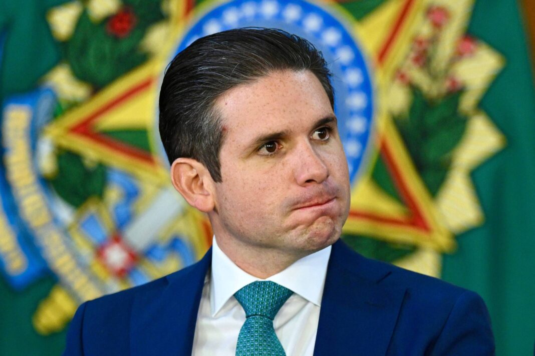 Um homem com cabelo liso e escuro, usando um terno azul e uma gravata verde, está sentado em um evento oficial. Ele tem uma expressão pensativa, com os lábios comprimidos. Ao fundo, há um emblema verde e dourado, que parece ser um símbolo nacional ou institucional.