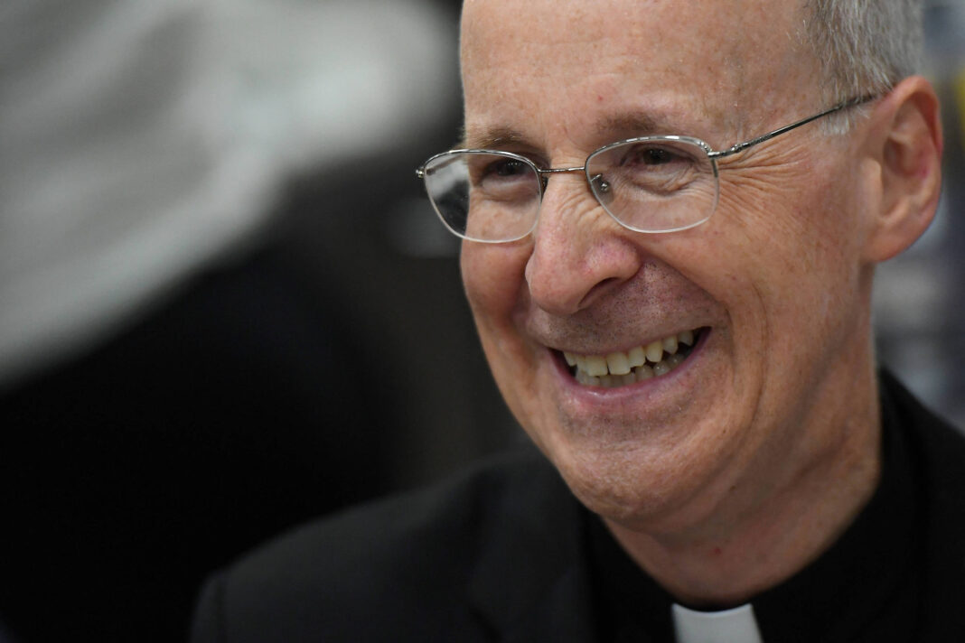 Um homem de meia-idade, com cabelo grisalho e óculos, está sorrindo. Ele usa uma roupa preta com uma colarinho clerical branco. O fundo é desfocado, sugerindo um ambiente de evento ou reunião.