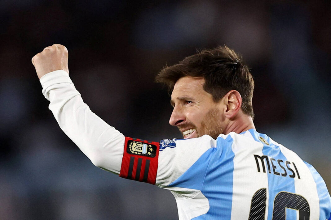 Um jogador de futebol está levantando o braço em sinal de comemoração. Ele usa uma camisa de futebol com listras verticais em azul e branco, e tem um distintivo de capitão na manga. O jogador tem cabelo castanho e está sorrindo, com um fundo desfocado que sugere um estádio.