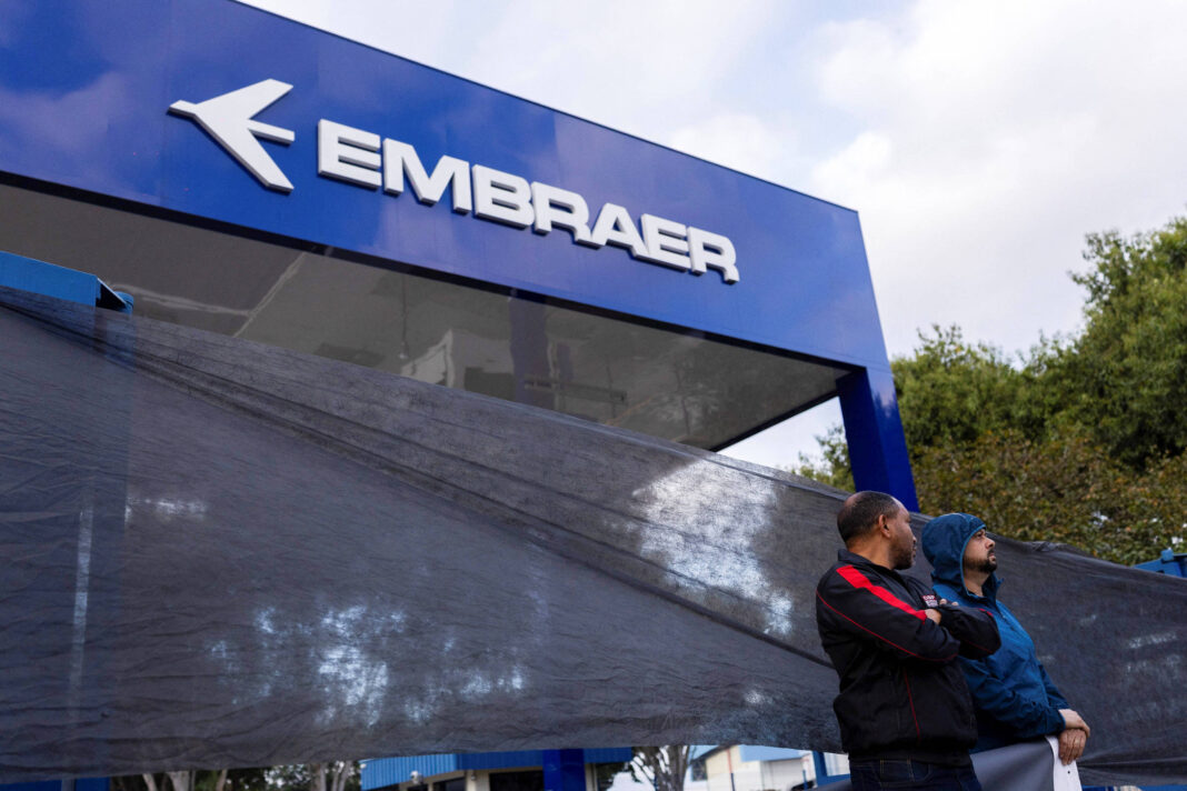 A imagem mostra a fachada da empresa Embraer, com um grande letreiro azul e o logotipo da empresa em destaque. Duas pessoas estão em pé em frente ao letreiro, uma delas vestindo um casaco escuro e a outra um casaco azul. Ao fundo, há uma área verde com árvores e um céu nublado.