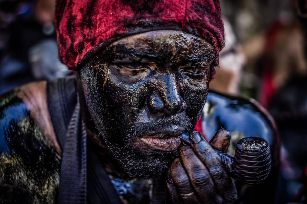 Pessoa com rosto e corpo cobertos por lama preta, usando um gorro vermelho. A pessoa segura um cachimbo na mão direita próxima à boca. Fundo desfocado com outras pessoas e elementos indistintos.