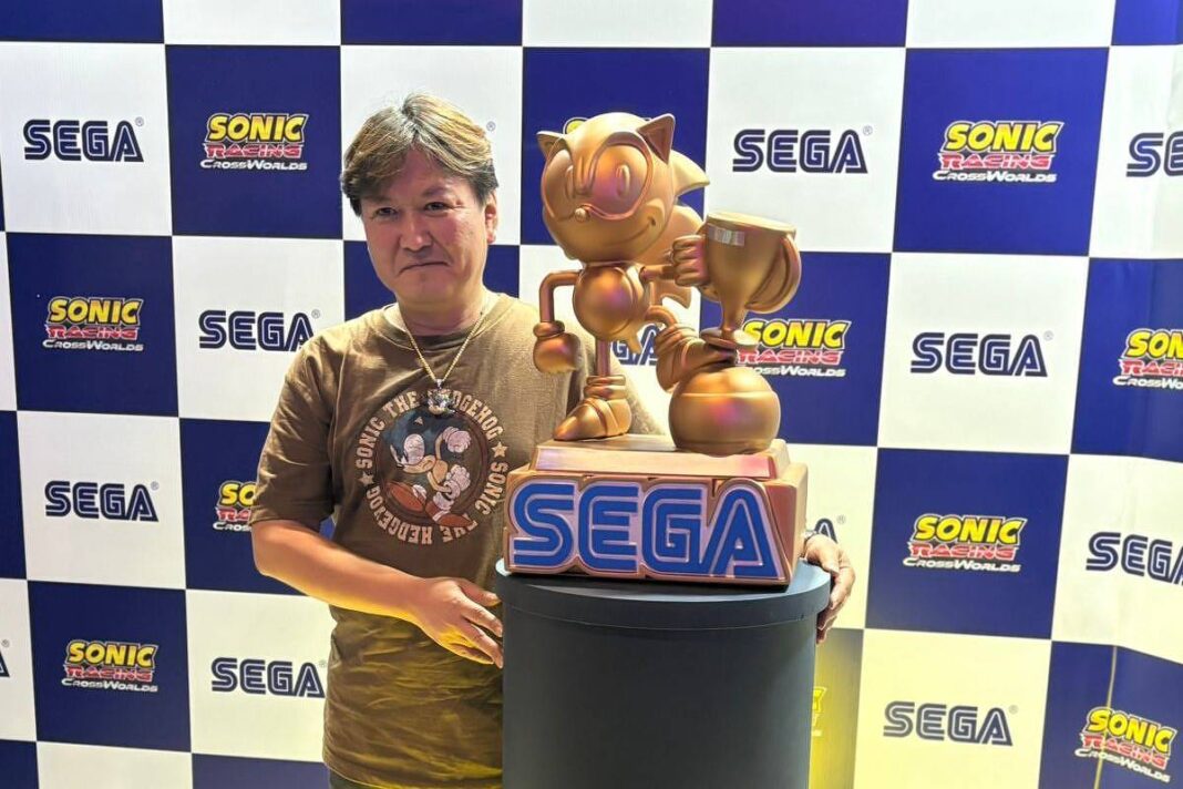Homem de camiseta marrom e calça amarela está ao lado de estátua dourada do personagem Sonic sobre pedestal iluminado com logo da SEGA. Fundo com painéis azuis e brancos exibindo logos da SEGA e Sonic.
