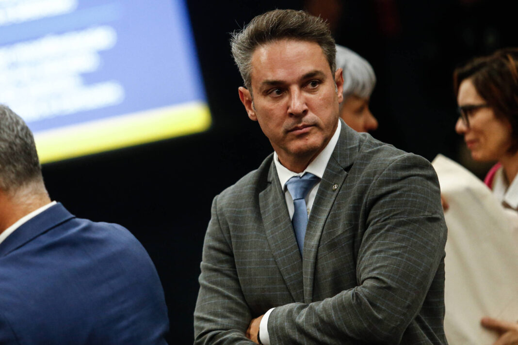 Um homem com cabelo curto e bem arrumado, vestindo um terno cinza e uma gravata azul, está em pé em um ambiente que parece ser um evento formal. Ele tem uma expressão séria e está olhando para o lado, enquanto outras pessoas estão ao fundo, algumas delas conversando. O fundo é desfocado, mas há uma tela visível com texto.
