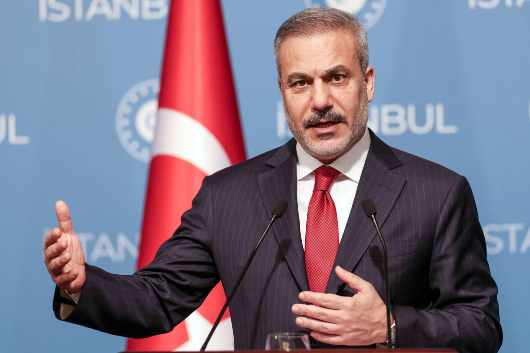 Homem de meia-idade com cabelo grisalho e barba curta veste terno escuro e gravata vermelha, falando em púlpito com dois microfones. Ao fundo, bandeira da Turquia e painel azul com texto 