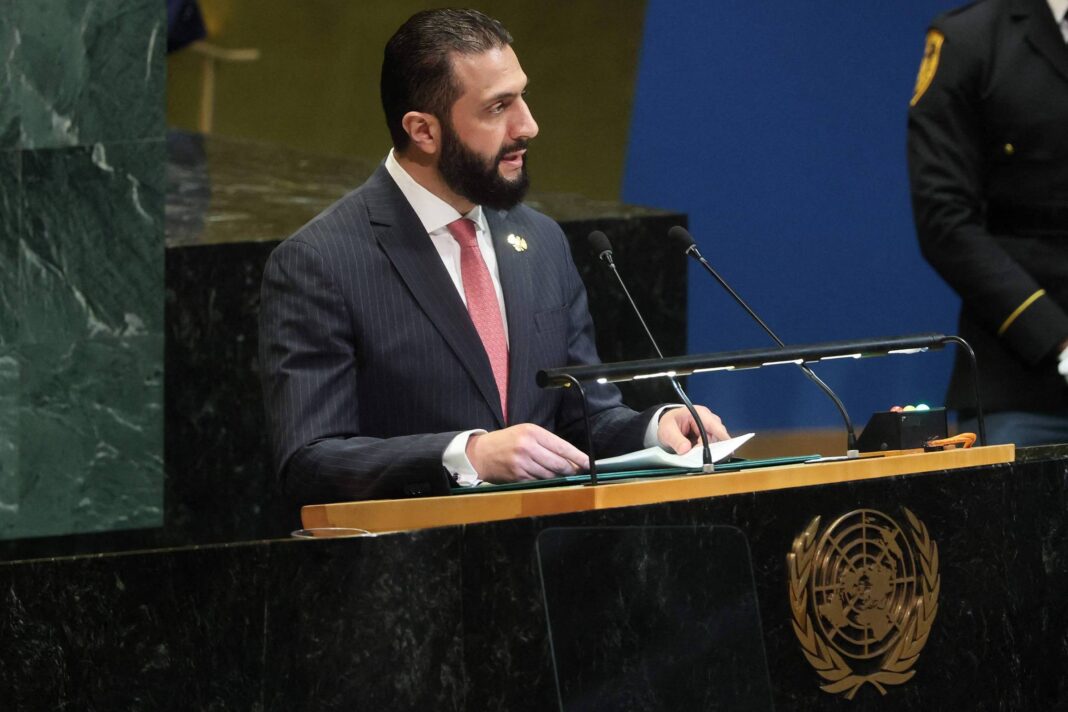 Homem de terno escuro e gravata rosa fala no púlpito da Assembleia Geral da ONU, com o emblema da organização visível. Um policial em uniforme formal está em pé ao fundo, à direita.