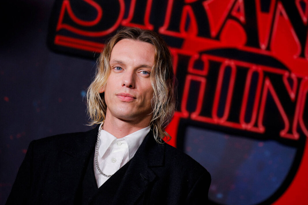 Homem de cabelos loiros e ondulados veste terno preto com camisa branca e corrente prateada, posando diante de logo vermelho e preto de Stranger Things.