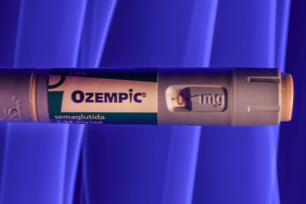 A imagem mostra uma caneta de medicamento chamada Ozempic, que contém semaglutida. A caneta é de cor cinza com uma etiqueta verde e branca, onde está escrito 