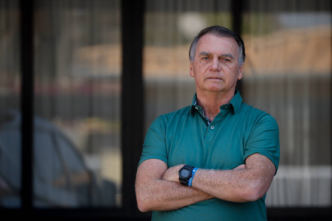 Um homem está em pé com os braços cruzados, olhando para a câmera. Ele usa uma camisa verde e tem cabelo curto e grisalho. Ao fundo, há uma janela com cortinas e uma parede. A iluminação é natural, sugerindo que a foto foi tirada durante o dia.