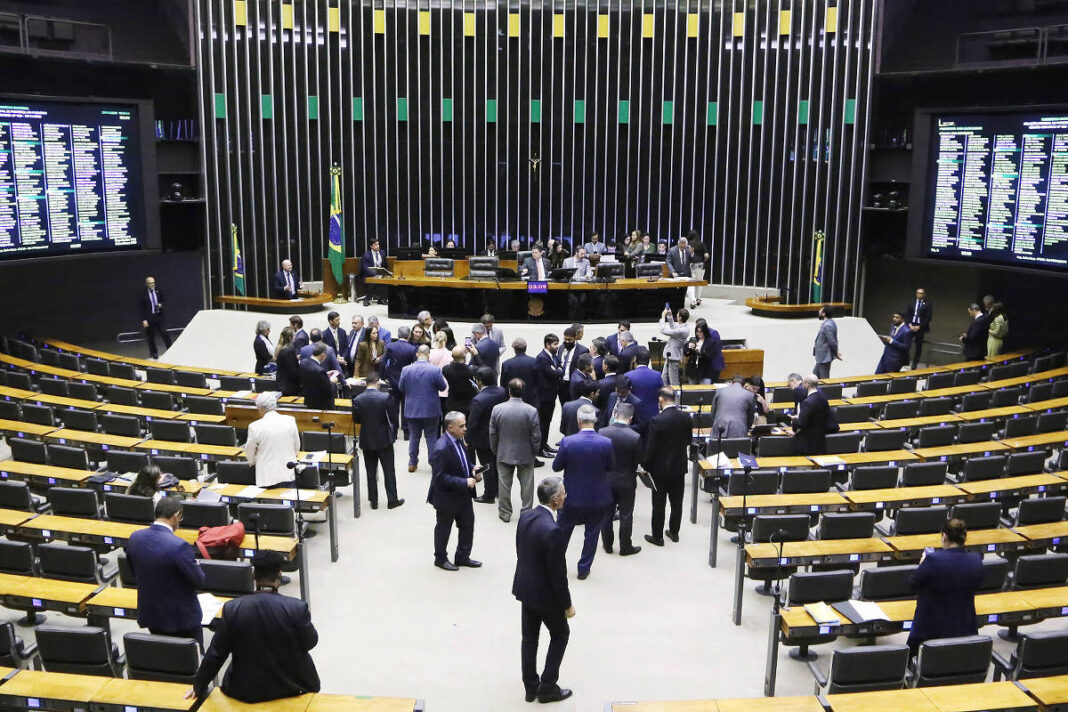Deputados reunidos no plenário da Câmara dos Deputados, com alguns em pé conversando no centro e outros sentados nas cadeiras amarelas. Painéis eletrônicos exibem informações legislativas nas laterais e ao fundo, atrás da mesa principal, há a bandeira do Brasil.