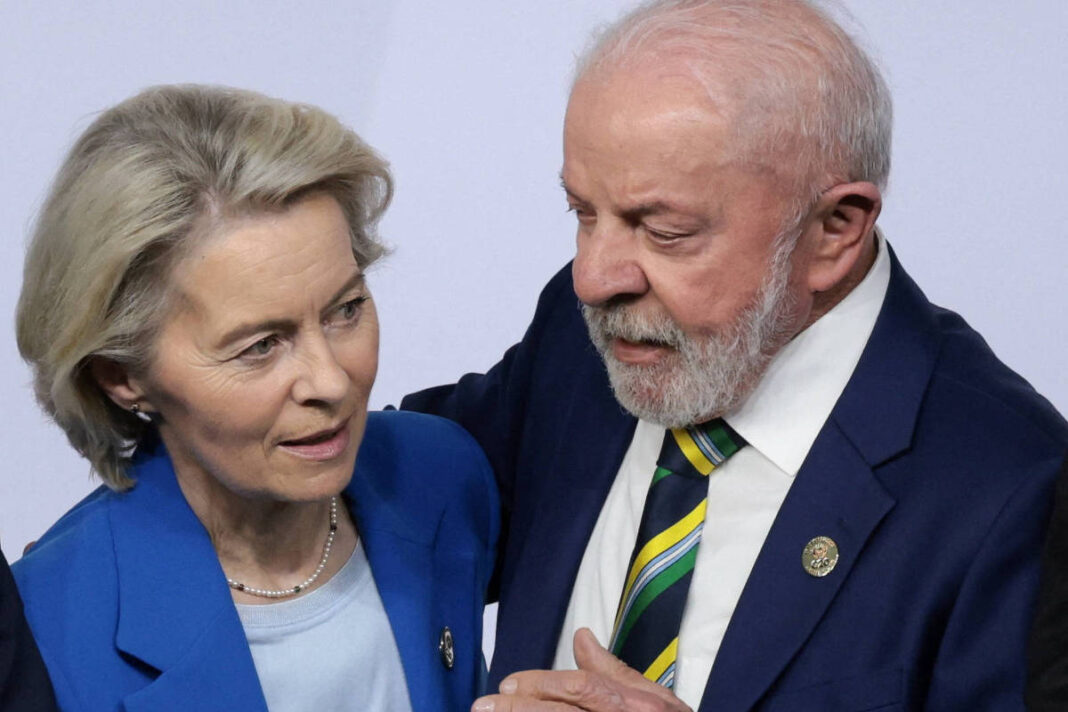 Homem e mulher em trajes formais conversam próximos um do outro. Homem usa terno azul escuro com gravata listrada verde, amarela e preta; mulher veste blazer azul e blusa clara. Fundo neutro claro.