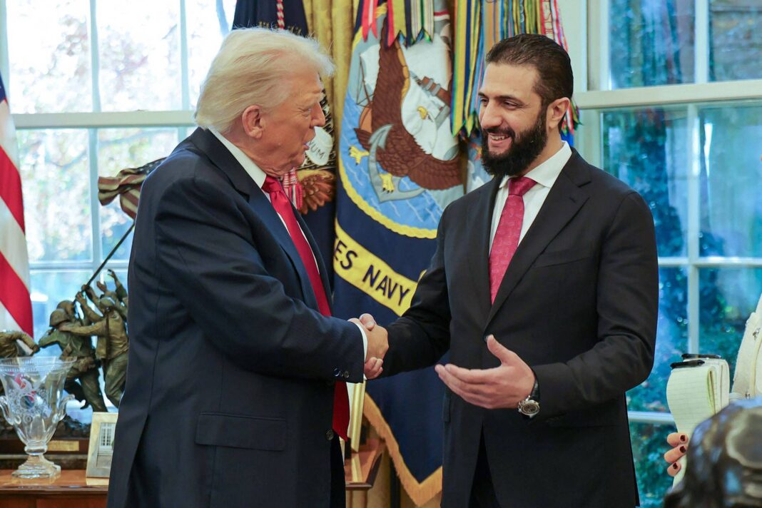 Dois homens de terno apertam as mãos em sala com bandeiras e decoração oficial ao fundo. Um deles tem cabelo branco e o outro barba e cabelo escuros.