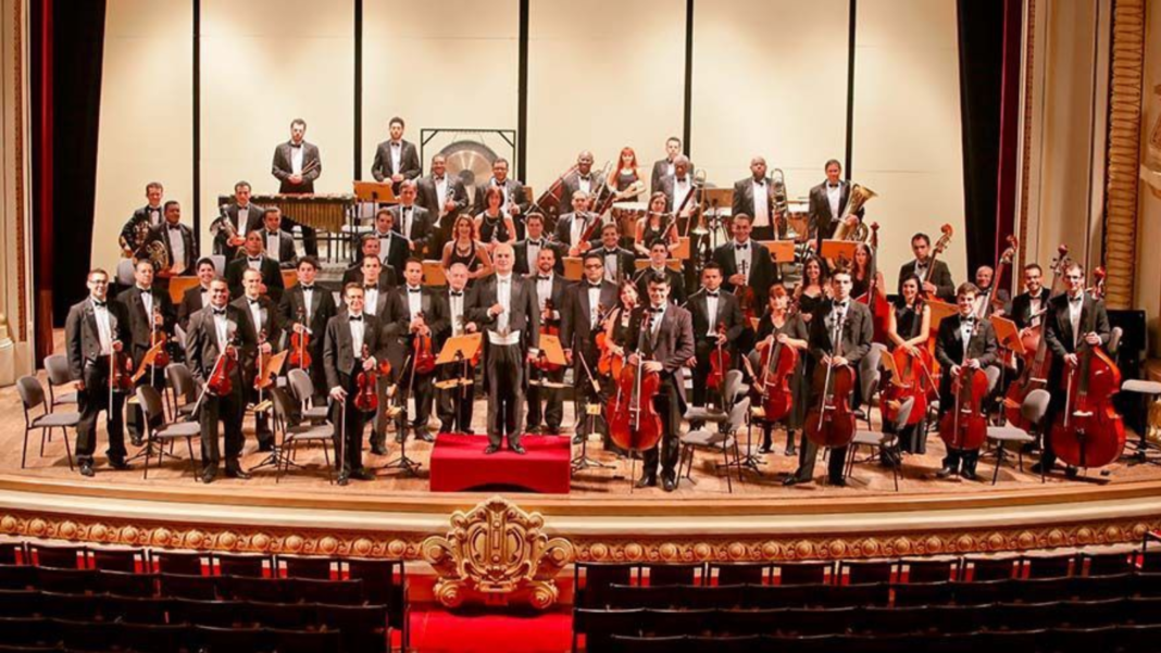 Orquestra sinfônica reunida no palco de um teatro histórico, com músicos e maestro em formação clássica, simbolizando a força da música como expressão do Espírito do Natal e da comunhão humana.
