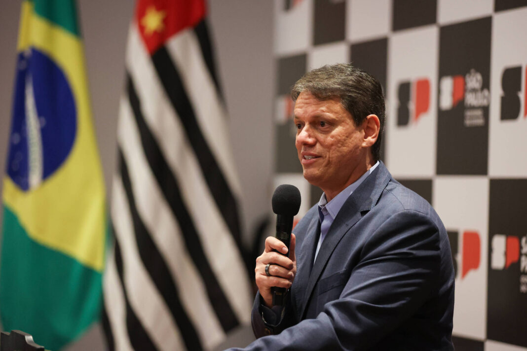 Homem de terno azul fala ao microfone em evento com bandeiras do Brasil e de São Paulo ao fundo. Painel com logotipo do governo paulista aparece atrás.