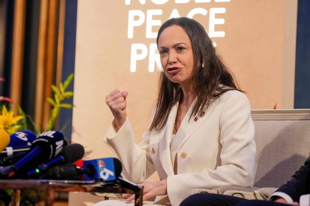 Mulher de cabelos longos e lisos, vestindo blazer branco, fala com expressão enfática em frente a um painel com o texto 