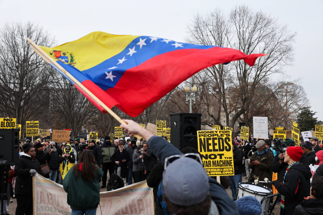 Pessoa segura bandeira da Venezuela em protesto ao ar livre com dezenas de manifestantes ao fundo. Muitos seguram cartazes amarelos com texto 