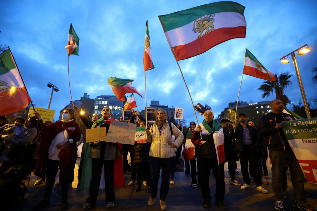 Grupo de manifestantes segura bandeiras da antiga bandeira do Irã e cartazes em praça urbana ao entardecer. Edifícios e postes de iluminação pública aparecem ao fundo sob céu azul com nuvens.