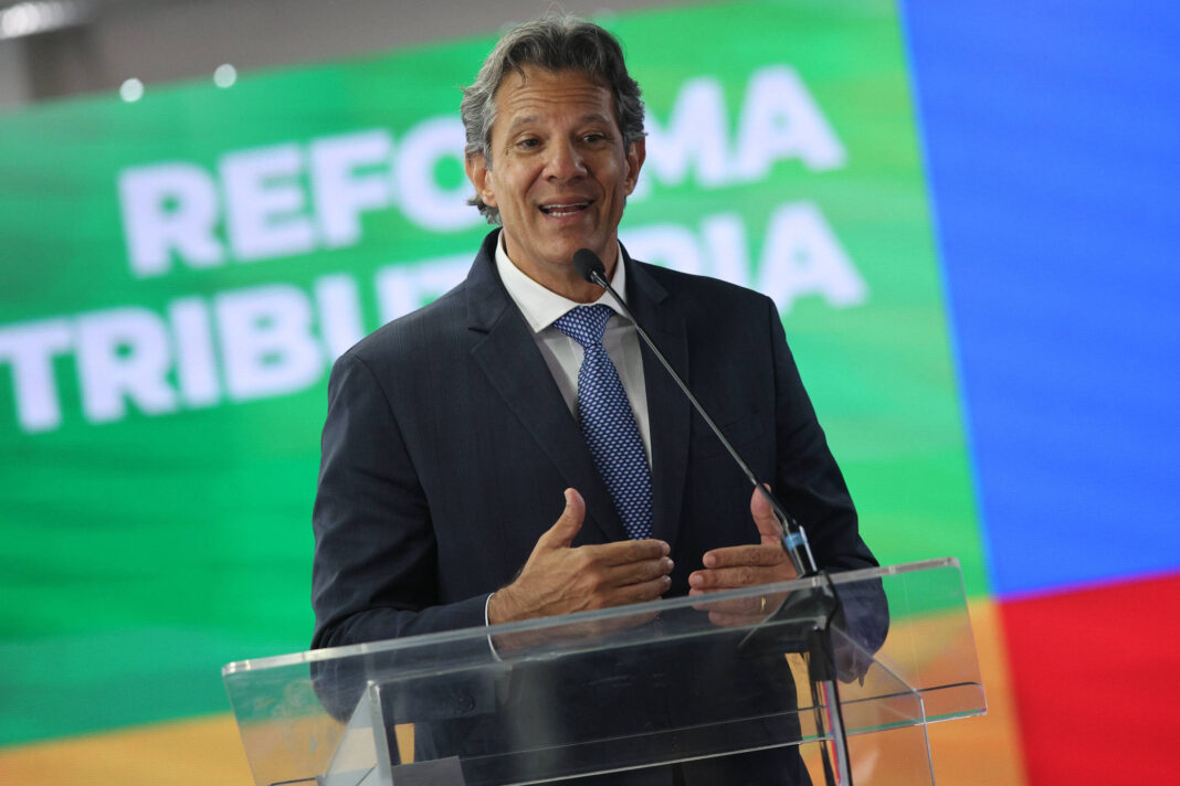 Homem de terno escuro e gravata azul fala em microfone atrás de púlpito transparente. Ao fundo, painel colorido exibe texto 