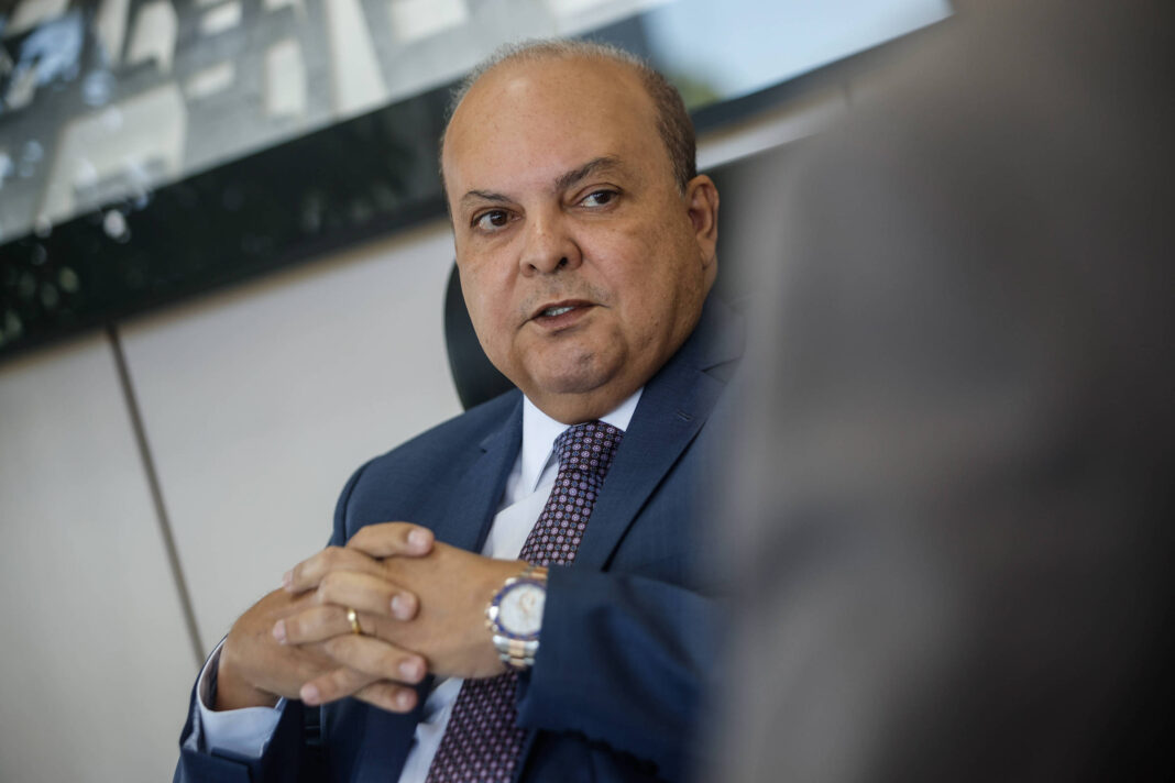 Um homem de pele clara, com cabelo curto e calvo, está sentado em uma cadeira. Ele usa um terno escuro e uma gravata roxa. Suas mãos estão unidas na frente dele. Ao fundo, há uma parede com uma imagem de uma cidade ou um projeto arquitetônico. A iluminação é suave e o ambiente parece ser um escritório.