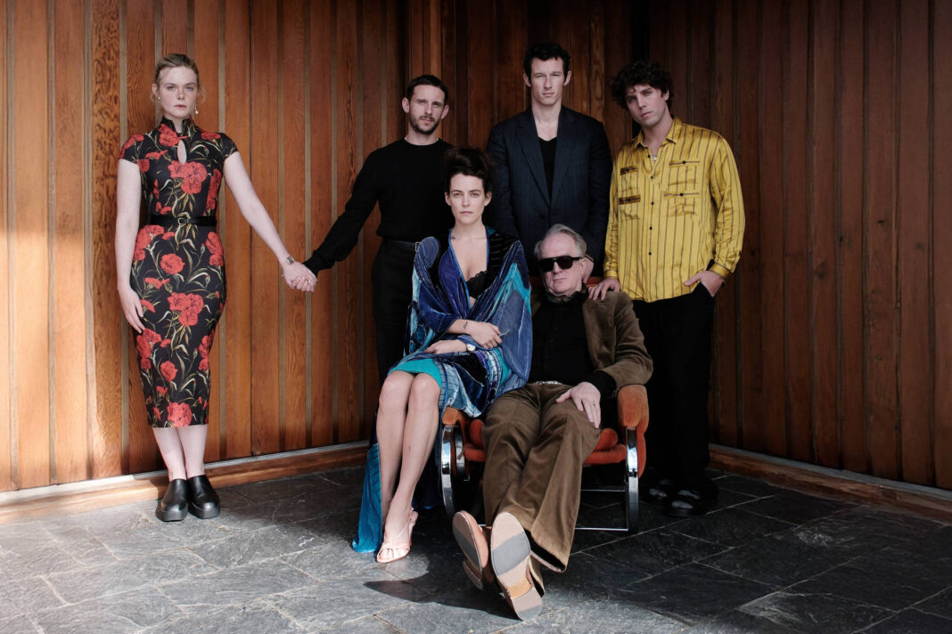 Cinco pessoas posam em um ambiente interno com paredes de madeira. Uma mulher de vestido floral vermelho e preto está em pé à esquerda, segurando a mão de um homem de camiseta preta. No centro, uma mulher sentada usa vestido azul e está ao lado de um homem idoso de óculos escuros e roupa marrom sentado em uma cadeira. À direita, dois homens estão em pé, um com camisa preta e outro com camisa amarela listrada.