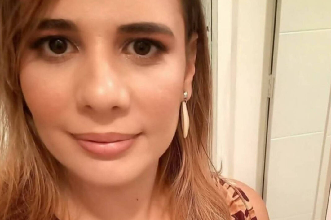 Mulher jovem com cabelo loiro e liso, usando brincos alongados e blusa estampada em tons de marrom e vinho, posando em ambiente interno com parede clara ao fundo.