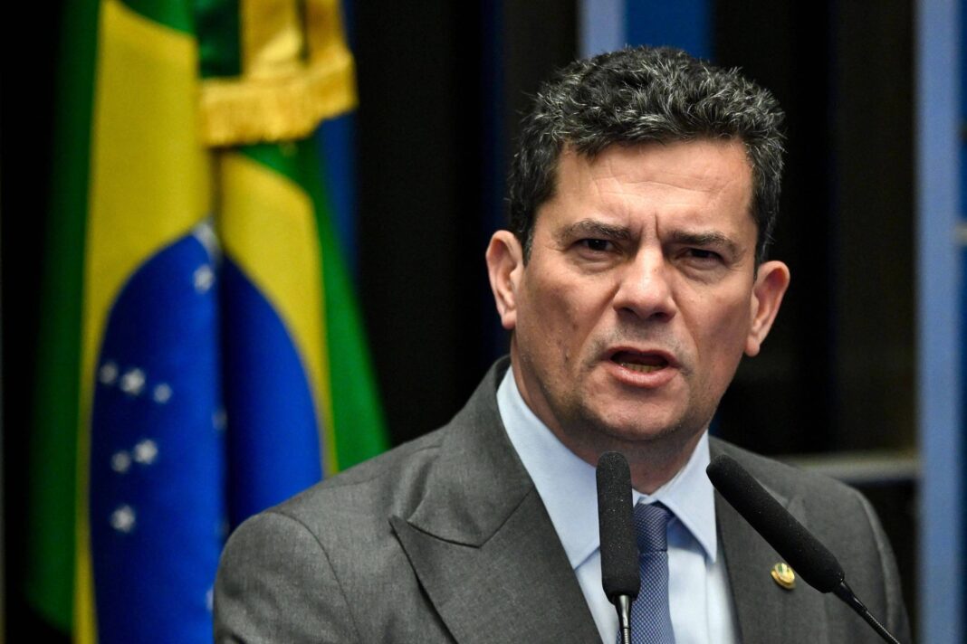 Homem de terno cinza e gravata azul fala em microfones diante de bandeira do Brasil desfocada ao fundo.