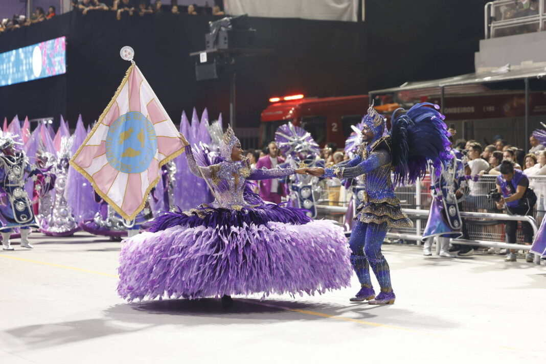 Casal com trajes elaborados em tons de roxo e azul dança em avenida durante desfile. Mulher segura bandeira rosa com círculo azul, vestido com saia volumosa de penas roxas. Homem usa fantasia com penas azuis e detalhes dourados. Público observa atrás de grades.