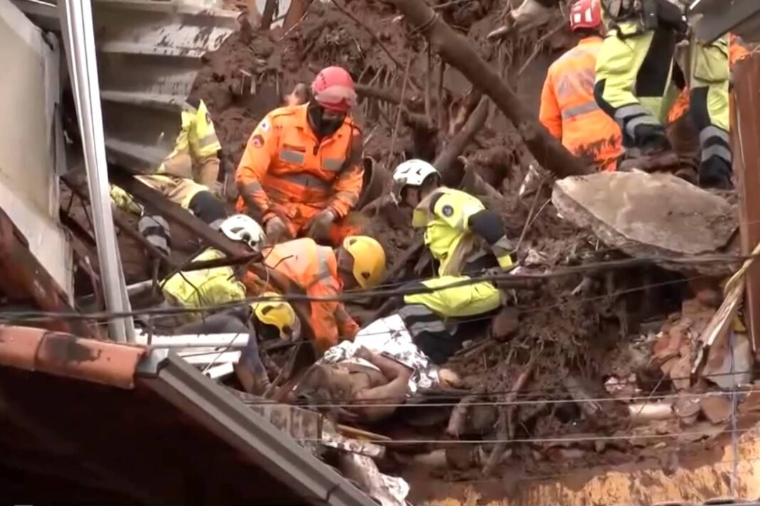 Seis socorristas com capacetes e roupas de proteção removem terra e escombros em área de desabamento. Estrutura danificada e cabos visíveis ao redor.