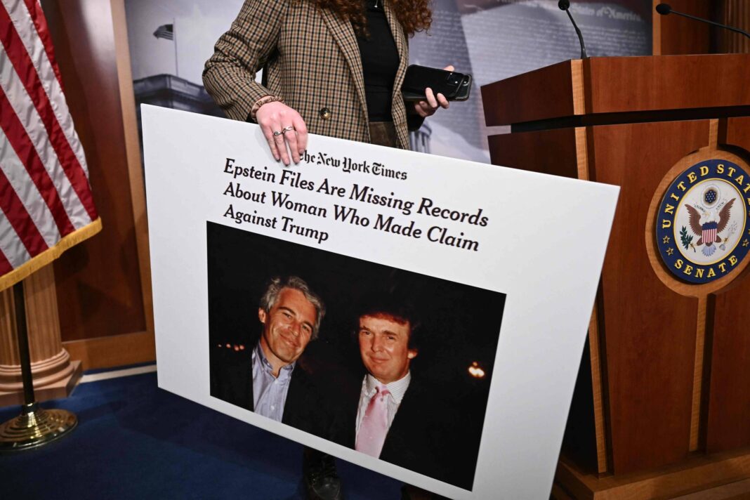 Mulher de cabelos cacheados e casaco xadrez segura cartaz grande com manchete do New York Times sobre arquivos Epstein e Trump. Cartaz mostra foto de Jeffrey Epstein e Donald Trump. Ao fundo, púlpito com selo do Senado dos EUA e duas bandeiras americanas.