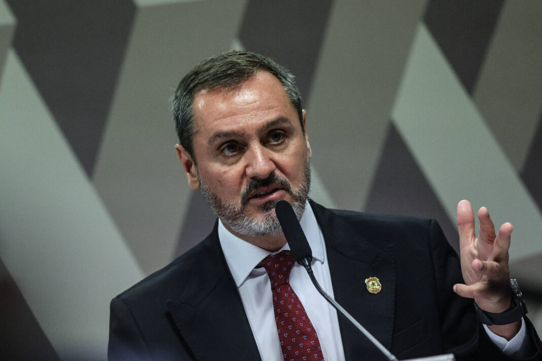 Homem de meia-idade com cabelo curto e barba grisalha veste terno preto, camisa branca e gravata vermelha, falando em microfone com mão direita levantada. Fundo com padrão geométrico em tons neutros.