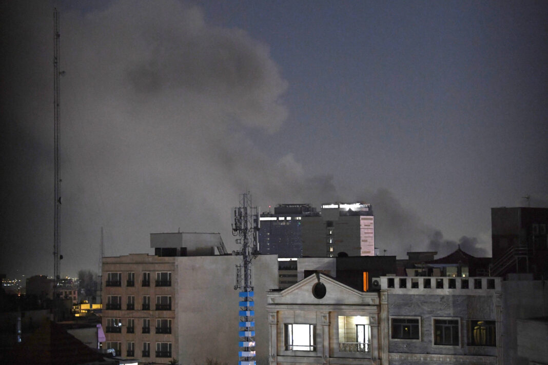 Fumaça escura e densa se eleva no céu noturno atrás de prédios residenciais e comerciais em área urbana. Algumas janelas estão iluminadas, e há torres de comunicação visíveis entre os edifícios.