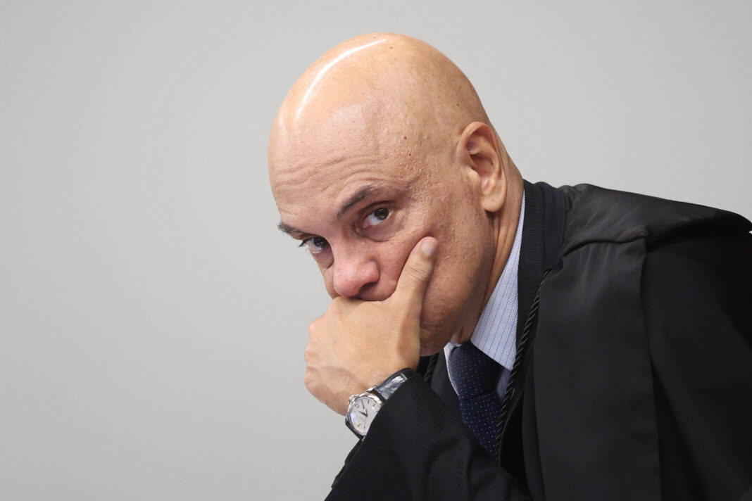 Homem calvo veste terno preto, camisa branca e gravata azul, apoiando o rosto na mão esquerda em gesto pensativo, com fundo cinza neutro.