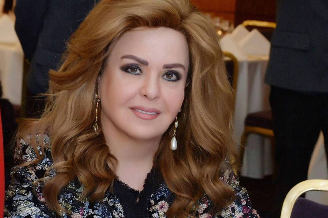 Mulher com cabelos loiros ondulados, maquiagem marcada e brincos longos, veste casaco floral escuro. Ela está sentada à mesa com copos de água e decoração floral branca à frente, em ambiente interno com outras pessoas ao fundo.