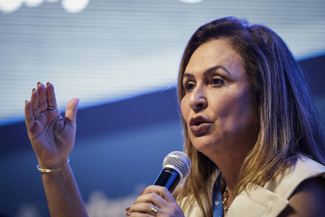 Uma mulher está falando em um microfone, levantando a mão direita em um gesto expressivo. Ela tem cabelo longo e solto, e está vestindo uma blusa clara. O fundo é desfocado, com uma iluminação suave e um padrão de luzes.