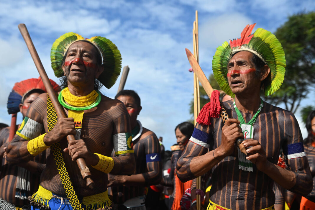 Homens indígenas com pintura corporal listrada preta e branca, cocares de penas verdes, amarelas e vermelhas, segurando bastões de madeira durante cerimônia ao ar livre sob céu azul.