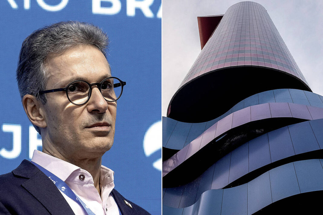 Homem de meia-idade com cabelo grisalho e óculos redondos veste camisa branca e paletó escuro, com fundo azul e texto parcialmente visível. Ao lado, vista de baixo para cima de edifício alto com fachada curva em tons de cinza e detalhes vermelhos, céu nublado ao fundo.