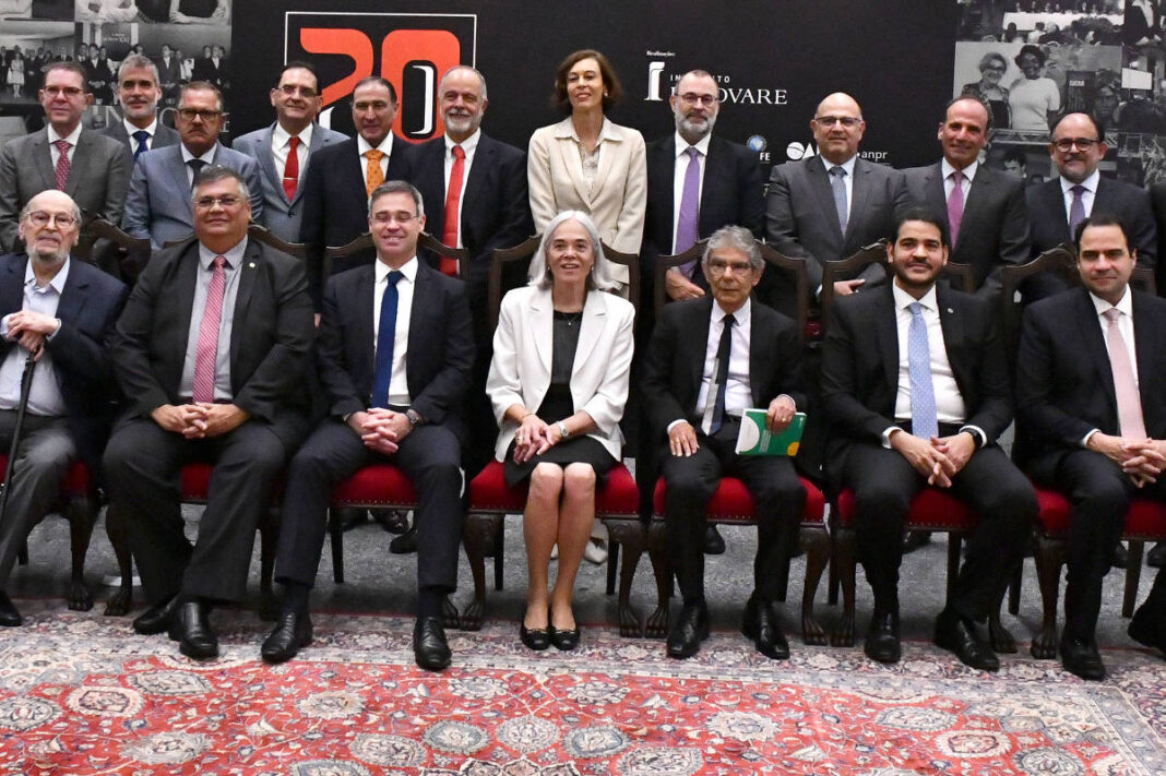 Vinte e quatro pessoas vestidas formalmente posam para foto oficial em ambiente interno com tapete persa no chão e painéis fotográficos ao fundo. Dezoito estão em pé e seis sentados em cadeiras vermelhas, todos olhando para a câmera.