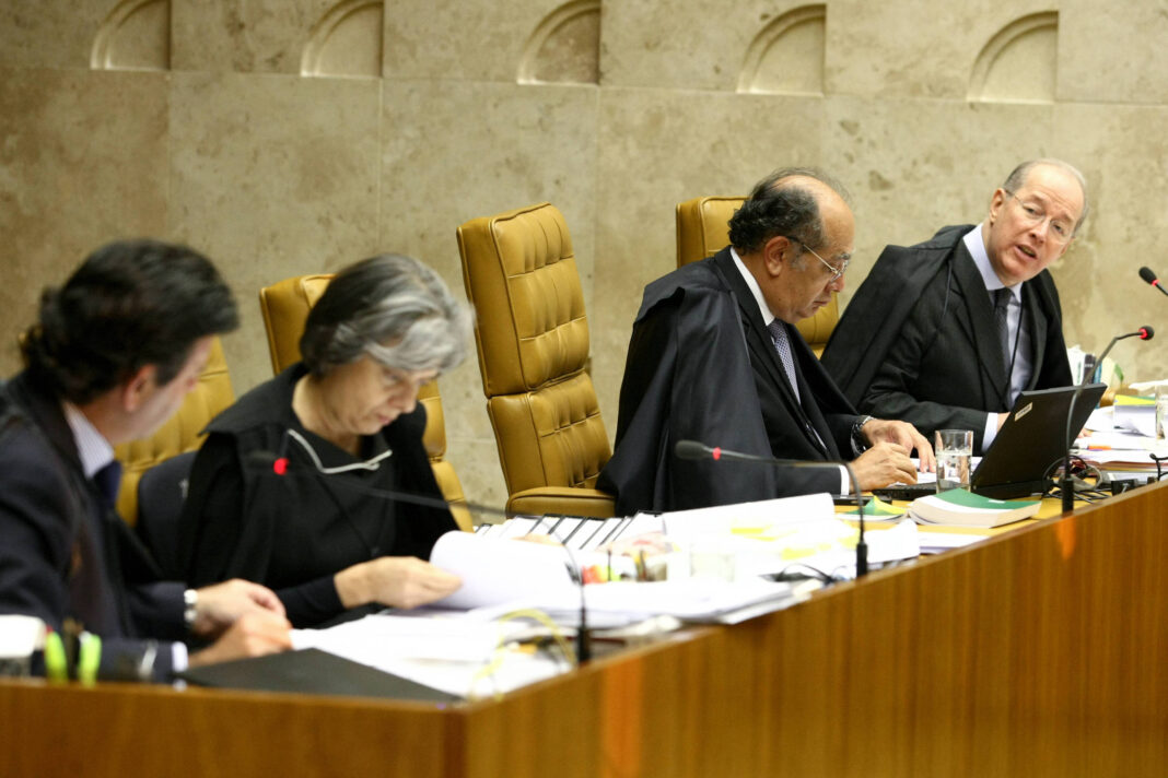Quatro juízes sentados em cadeiras altas de couro marrom em tribunal. Dois deles usam togas pretas, um está concentrado lendo documentos e outro fala olhando para o lado. Mesa à frente com microfones, papéis, livros e um laptop.