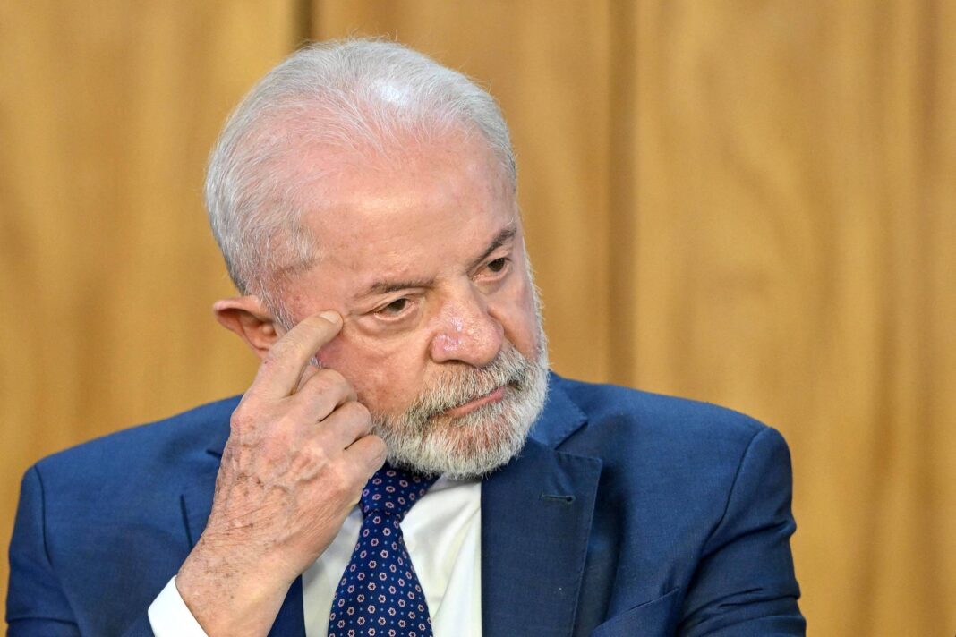 Um homem de cabelo grisalho e barba, vestindo um terno azul e uma gravata com padrões. Ele está sentado e parece estar pensativo, com a mão direita tocando o rosto, especificamente a área ao redor do olho. O fundo é de madeira clara, sugerindo um ambiente formal.