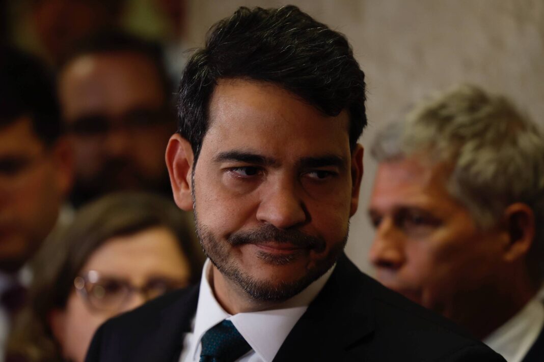 Homem de terno escuro e gravata verde escura olha para o lado em ambiente interno com outras pessoas desfocadas ao fundo.