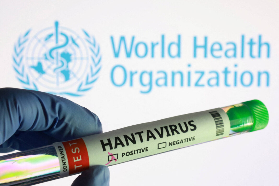 Mão com luva segura tubo de ensaio rotulado como teste de hantavírus com resultado positivo marcado. Ao fundo, logo da Organização Mundial da Saúde em azul.