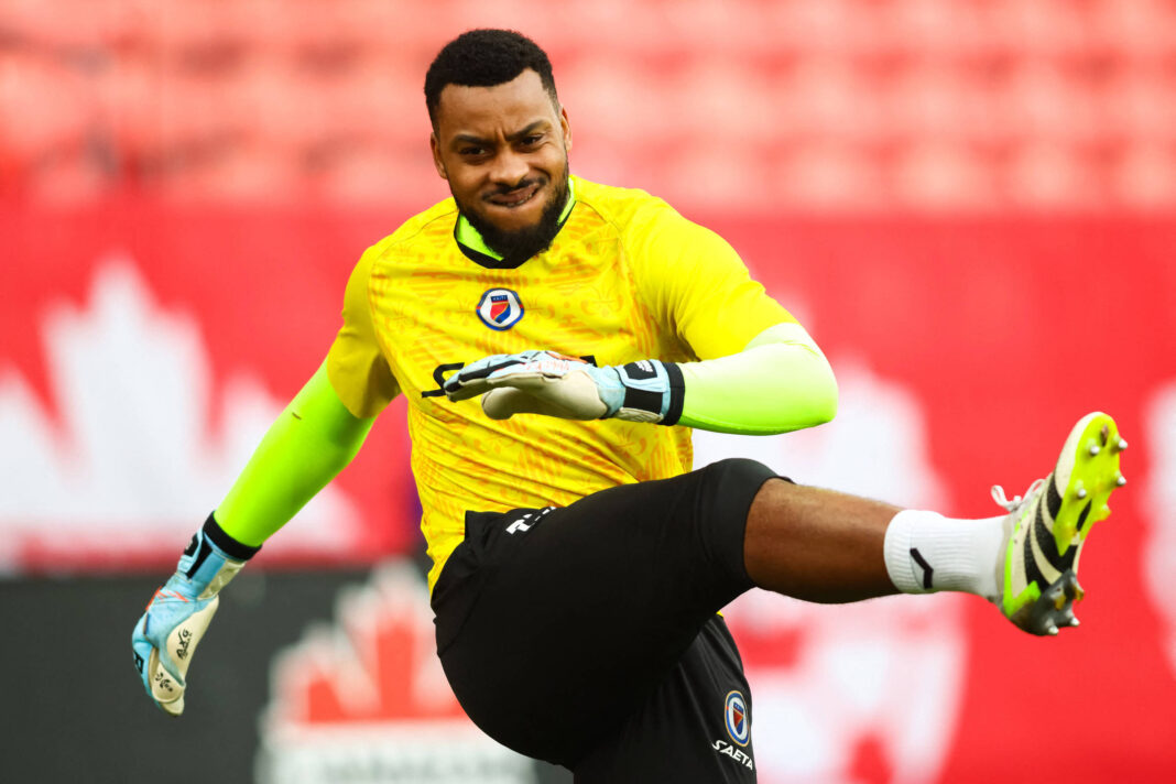 Goleiro com uniforme amarelo e calção preto chuta a bola durante treino em campo com arquibancada vermelha ao fundo.