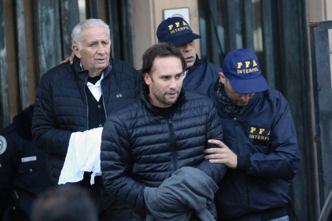 Homem de casaco preto é escoltado por dois agentes da Interpol com bonés azuis e jaquetas com a sigla PFA. Outro homem mais velho acompanha o grupo segurando um pano branco.