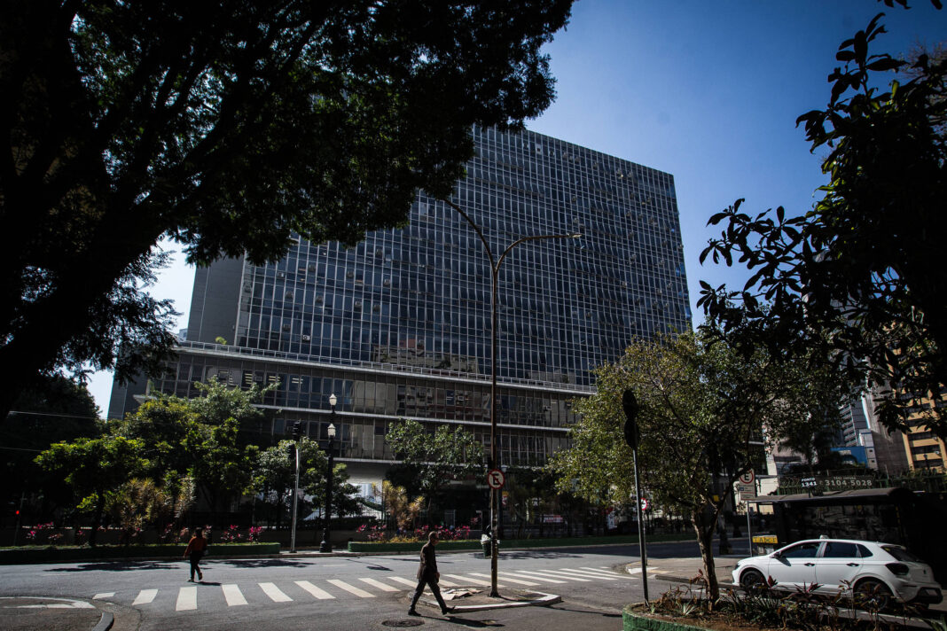 A imagem mostra um grande edifício de escritórios em São Paulo, com uma fachada de vidro refletindo o céu azul. Na parte inferior, há uma faixa de pedestres e algumas árvores ao redor. Algumas pessoas estão caminhando na calçada e um carro branco está estacionado próximo ao edifício.