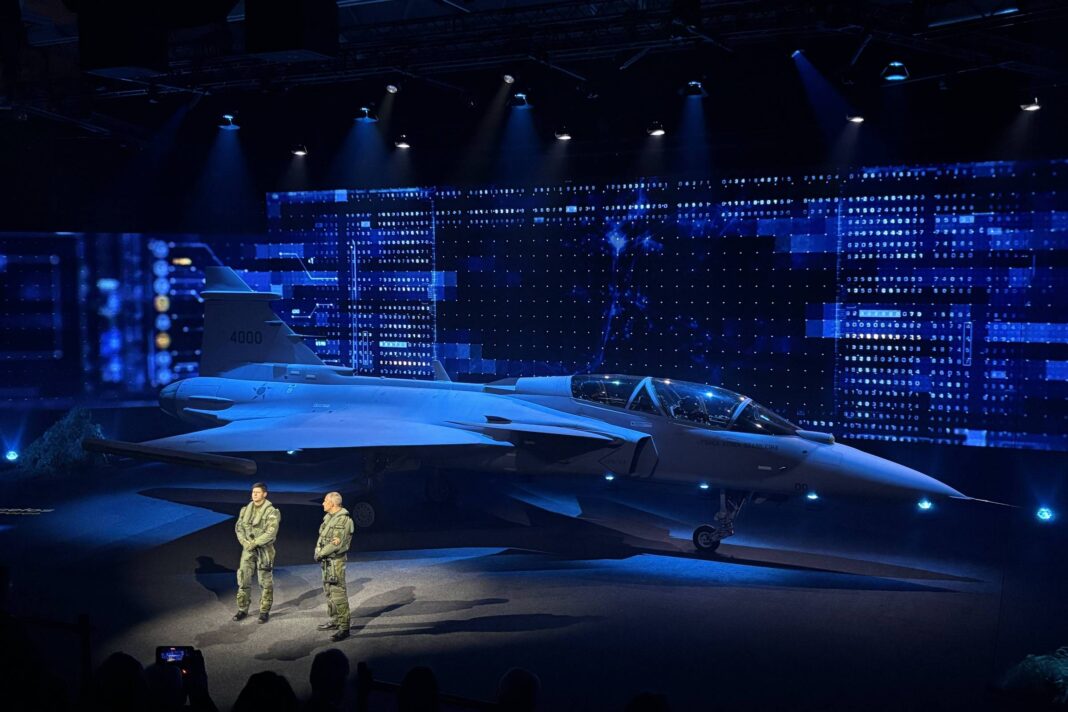 Caça militar cinza em exibição sob luzes azuis em palco escuro. Dois militares em uniforme verde estão em pé à frente da aeronave, com público em sombra na frente.