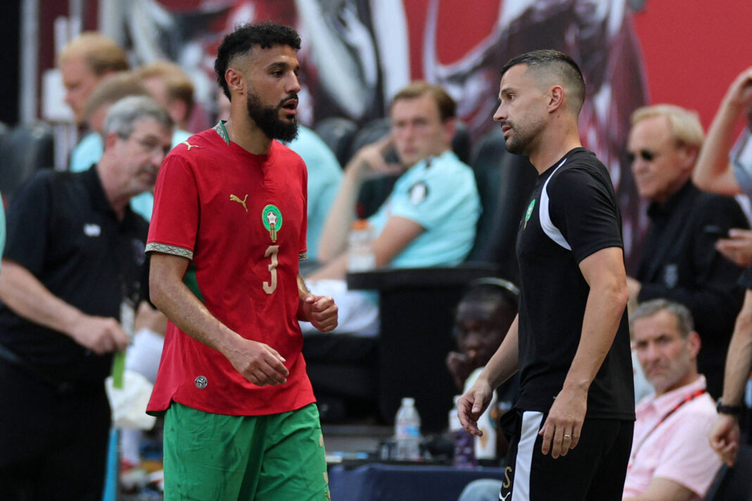Dois jogadores do Marrocos conversam à beira do campo durante partida. Um veste uniforme vermelho e verde com o número 3, o outro uniforme preto com detalhes brancos. Pessoas e equipe técnica aparecem ao fundo.